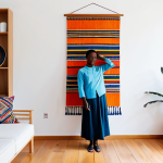 A professional woman, fully clothed in a modest, modern Scandinavian-style outfit, stands in a brightly lit, minimalist living room. A large, vibrant Imigongo-inspired textile wall hanging serves as a focal point, complemented by an intricately woven Agaseke basket on a nearby wooden shelf. The scene emphasizes the beautiful fusion of Rwandan cultural art with contemporary European interior design. perfect anatomy, correct proportions, natural pose, well-formed hands, proper finger count, natural body proportions, professional photography, high quality, safe for work, appropriate content, fully clothed, modest, family-friendly.